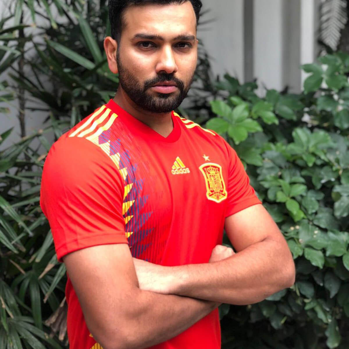Rohit Sharma 桌布 壁紙 — 一位留鬍子的男人自信地交叉雙臂站立，身穿紅色西班牙足球球衣，背景是茂密的綠色樹葉。