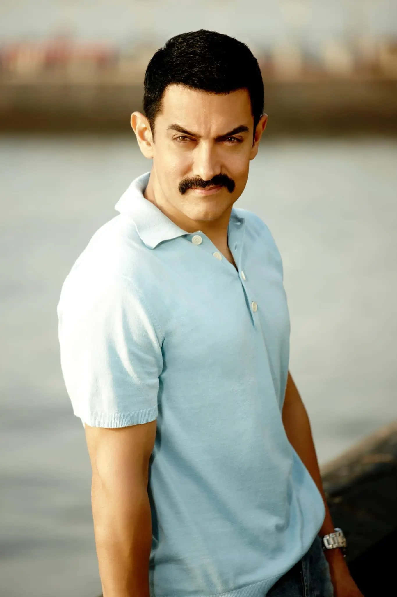 Aamir Khan 桌布 壁紙 — 一位留鬍子的男士穿著淺藍色Polo衫站在水邊，背景是模糊的城市天際線。