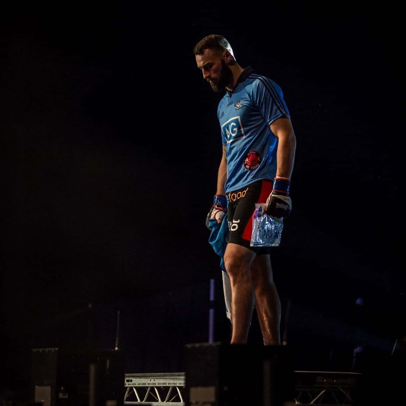 UFC 桌布 壁紙 — 一位留鬍子的男性運動員身穿藍色球衣和黑色短褲，獨自站在黑暗的舞台上，在戲劇性的聚光燈下拿著一個水瓶。