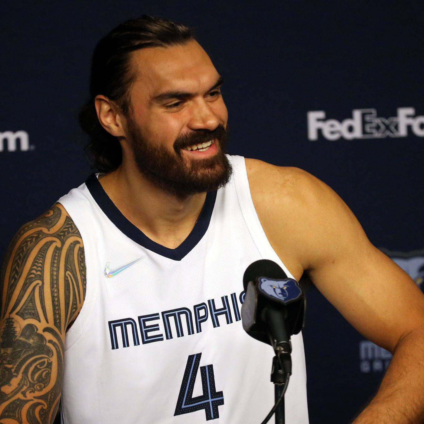 Steven Adams 桌布 壁紙 — 一位留鬍子的籃球運動員穿著曼菲斯灰熊隊球衣，在記者會上對著麥克風微笑說話。