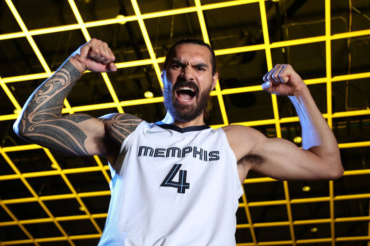 Steven Adams 桌布 壁紙 — 一位留鬍子的籃球運動員身穿曼菲斯球衣，對著發光的黃色網格天花板彎曲雙臂。