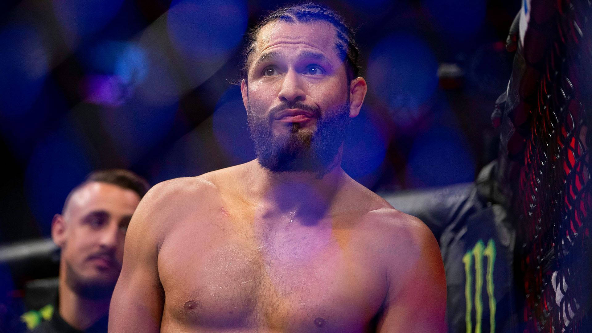 UFC 桌布 壁紙 — 一位留鬍子的綜合格鬥家在燈光昏暗的八角籠中，背景是模糊的觀眾和發光的燈光，眼神專注。