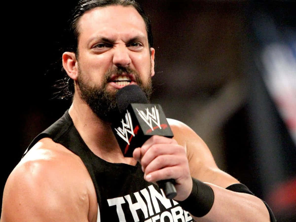 WWE 桌布 壁紙 — 一位留鬍子的肌肉摔角手穿著印有 'THINK BEFORE' 字樣的黑色背心，對著 WWE 麥克風大喊。
