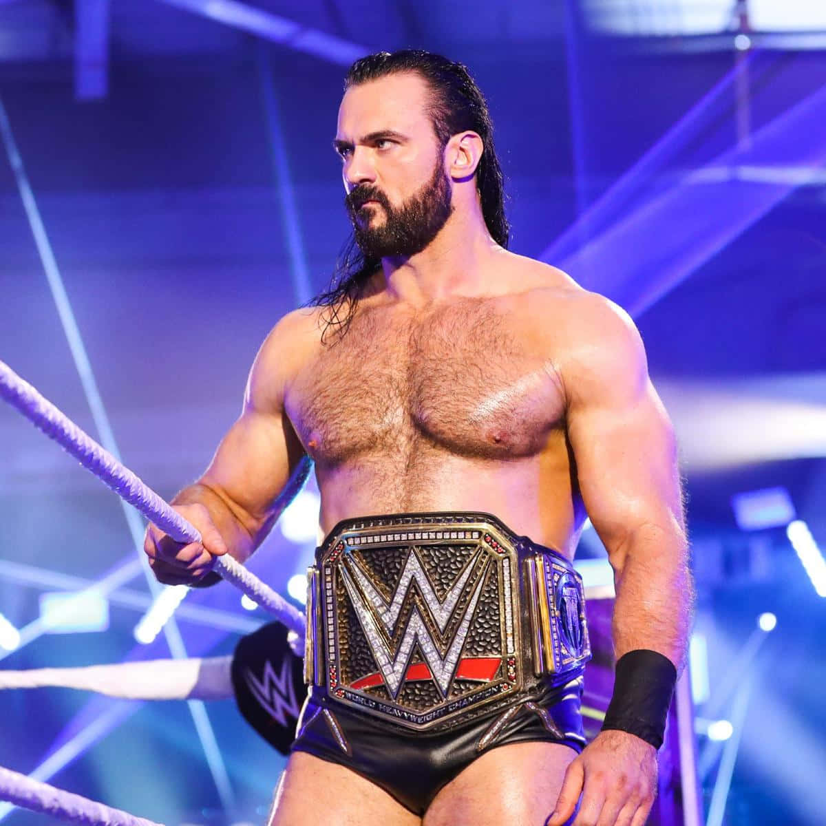 WWE 桌布 壁紙 — 一位留鬍子的肌肉發達摔角手站在擂台上，在藍色舞台燈光下高舉WWE冠軍腰帶。