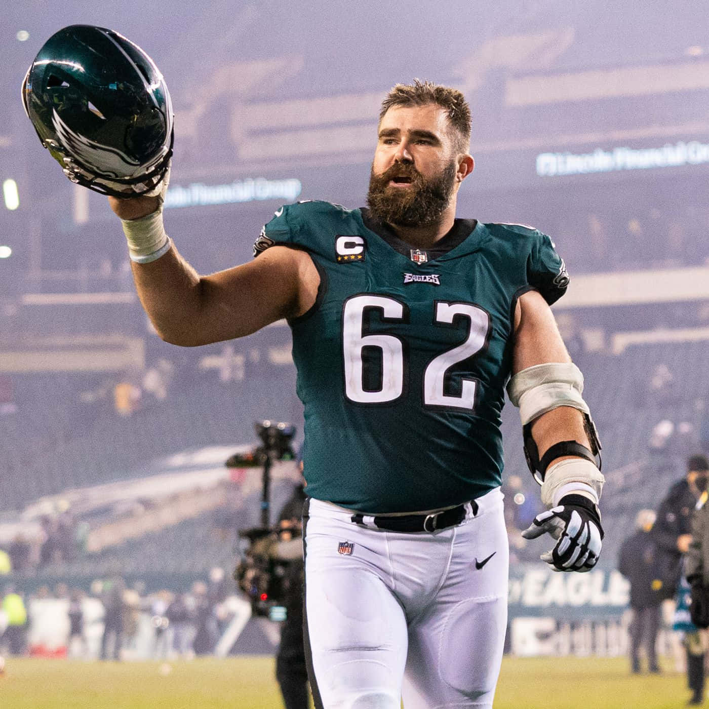 Jason Kelce 桌布 壁紙 — 一位留鬍子的費城老鷹隊美式足球員，身穿綠色62號球衣，在夜間球場上向球迷揮舞頭盔。