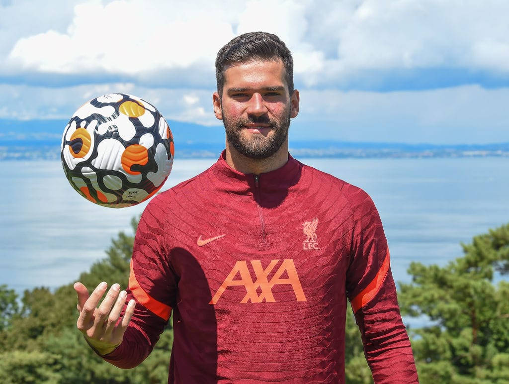 Alisson Becker 桌布 壁紙 — 一位留鬍子的足球運動員穿著紅色訓練服，在部分多雲的天空下，以藍色水和遠山為背景，平衡一個色彩繽紛的足球。