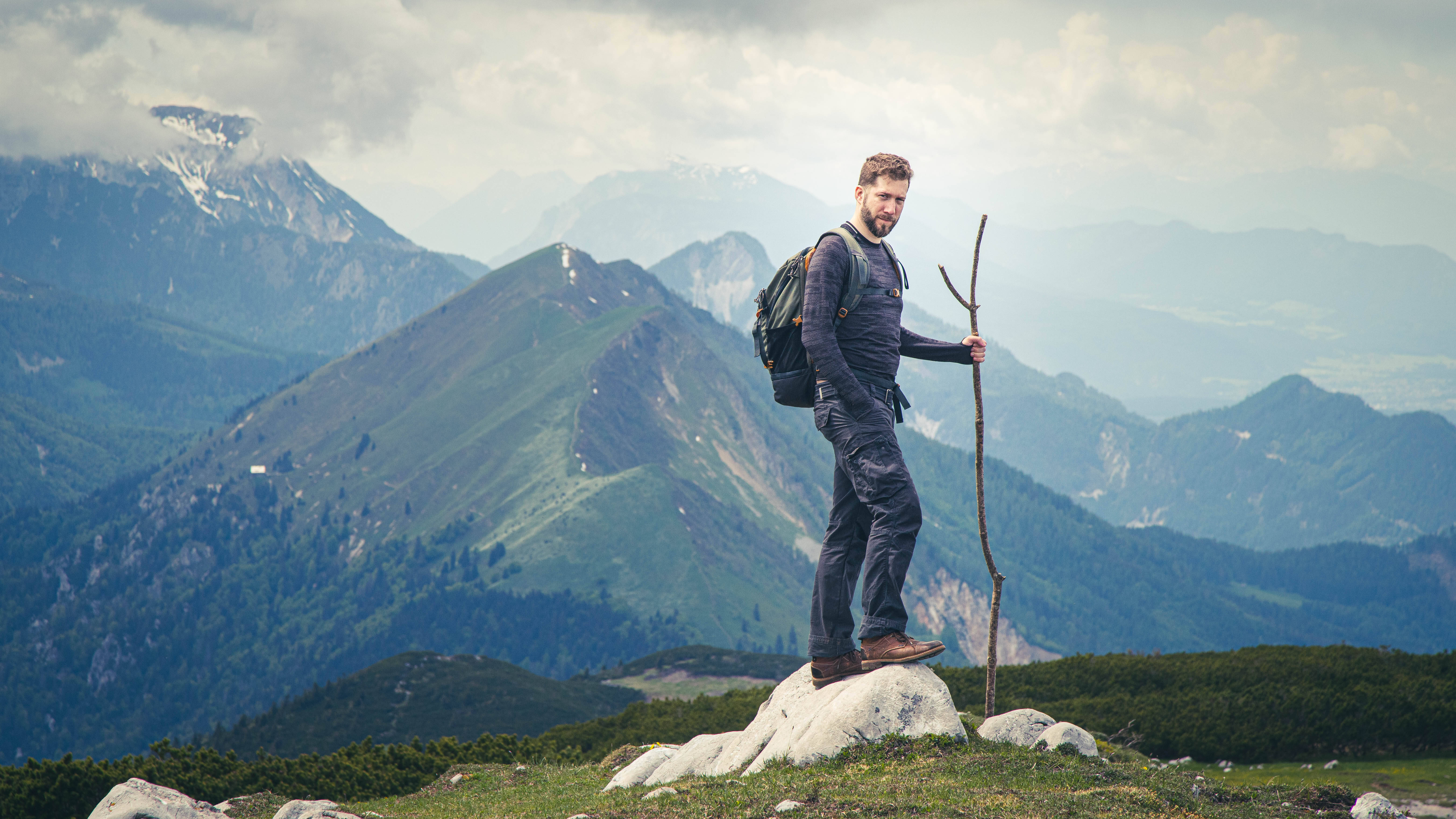 Slovenia 桌布 壁紙 — 一位留鬍子的遠足者站在岩石山頂上，手持登山杖，遠處是白雪覆蓋的山峰和森林覆蓋的山坡。