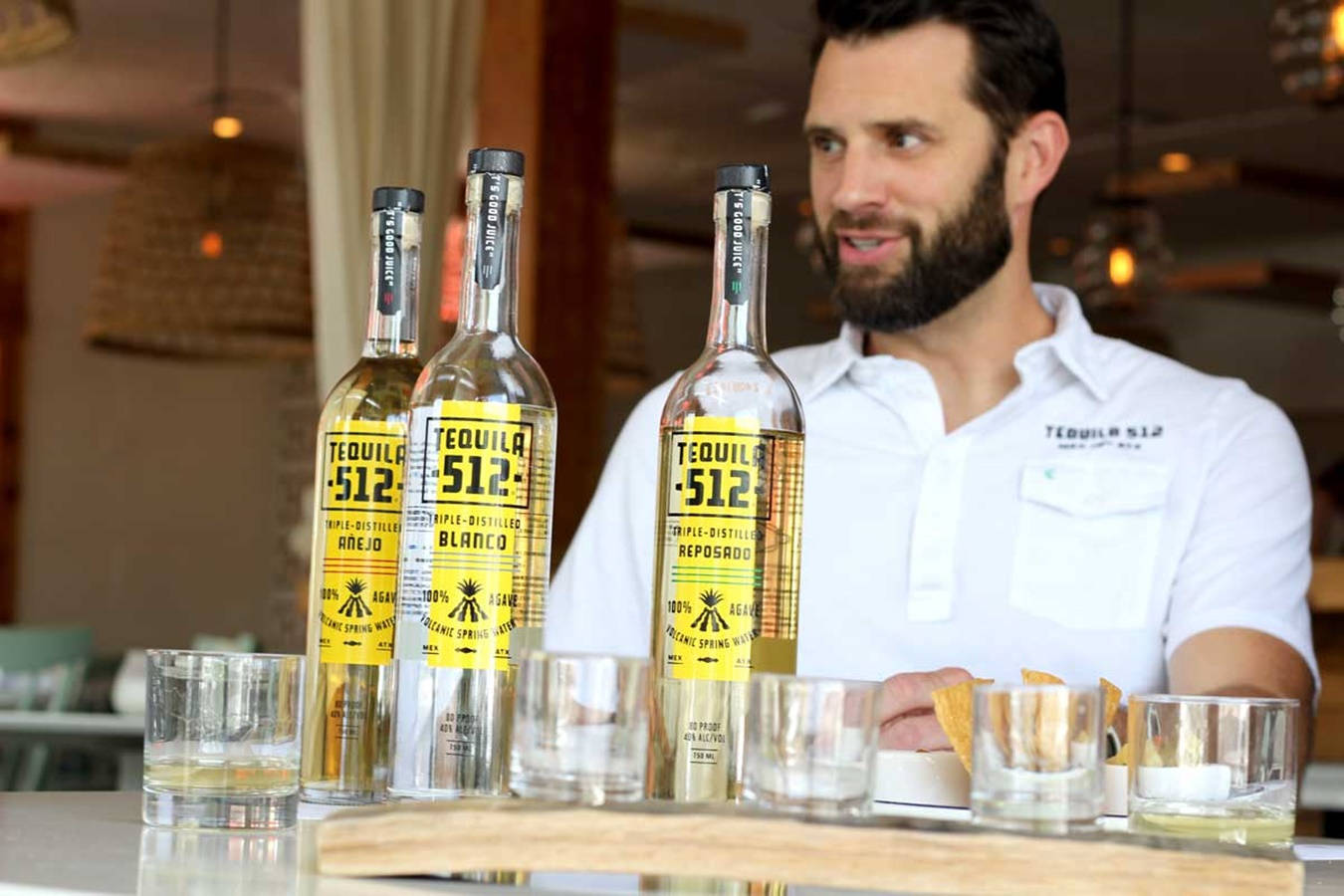 drinks 桌布 壁紙 — 一位留鬍子的酒保在吧檯後微笑，展示三瓶Tequila 512，搭配酒杯和玉米片。