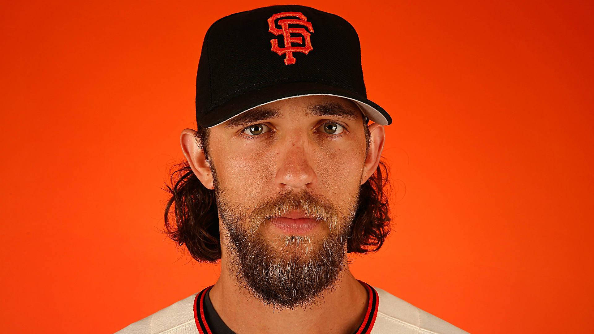 madison bumgarner 桌布 壁紙 — 一位留鬍子長髮的棒球運動員，戴著黑色帽子，上面有紅色SF標誌，背景為純橙色。