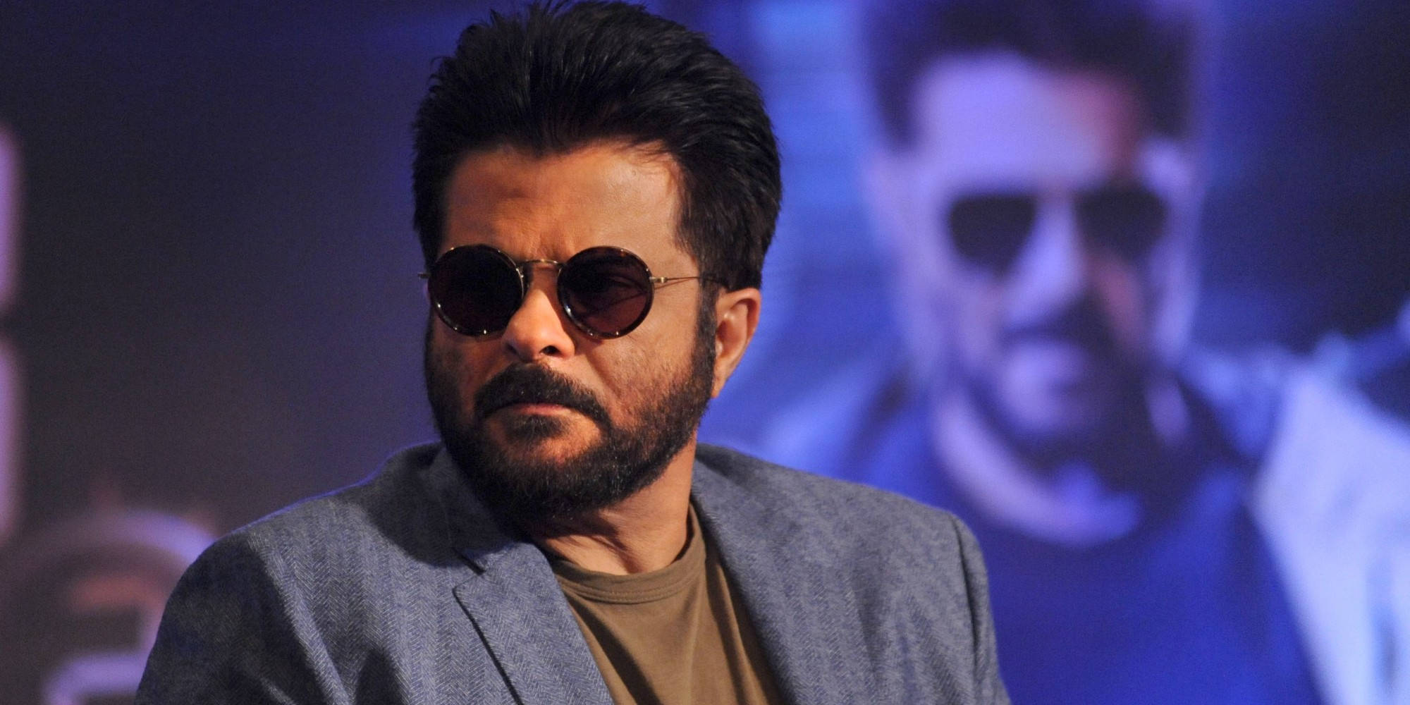 anil kapoor 桌布 壁紙 — 一位留鬍鬚、戴圓形太陽眼鏡的男子，穿著灰色西裝外套，背景是模糊的藍色。