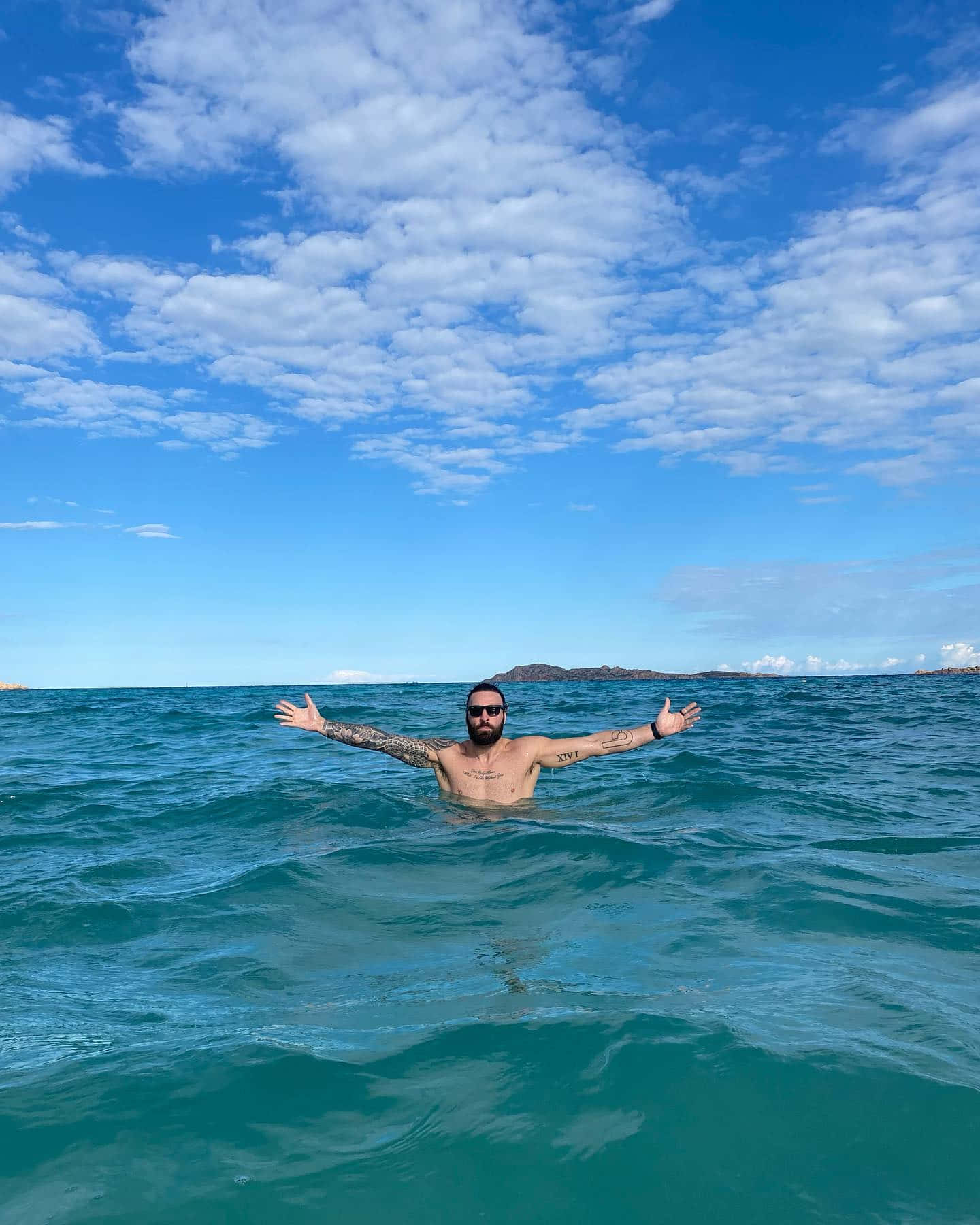 Vacation 桌布 壁紙 — 一位留鬍鬚、戴太陽眼鏡、有刺青的男人，在蔚藍天空和零星白雲下，於碧綠海水中張開雙臂。