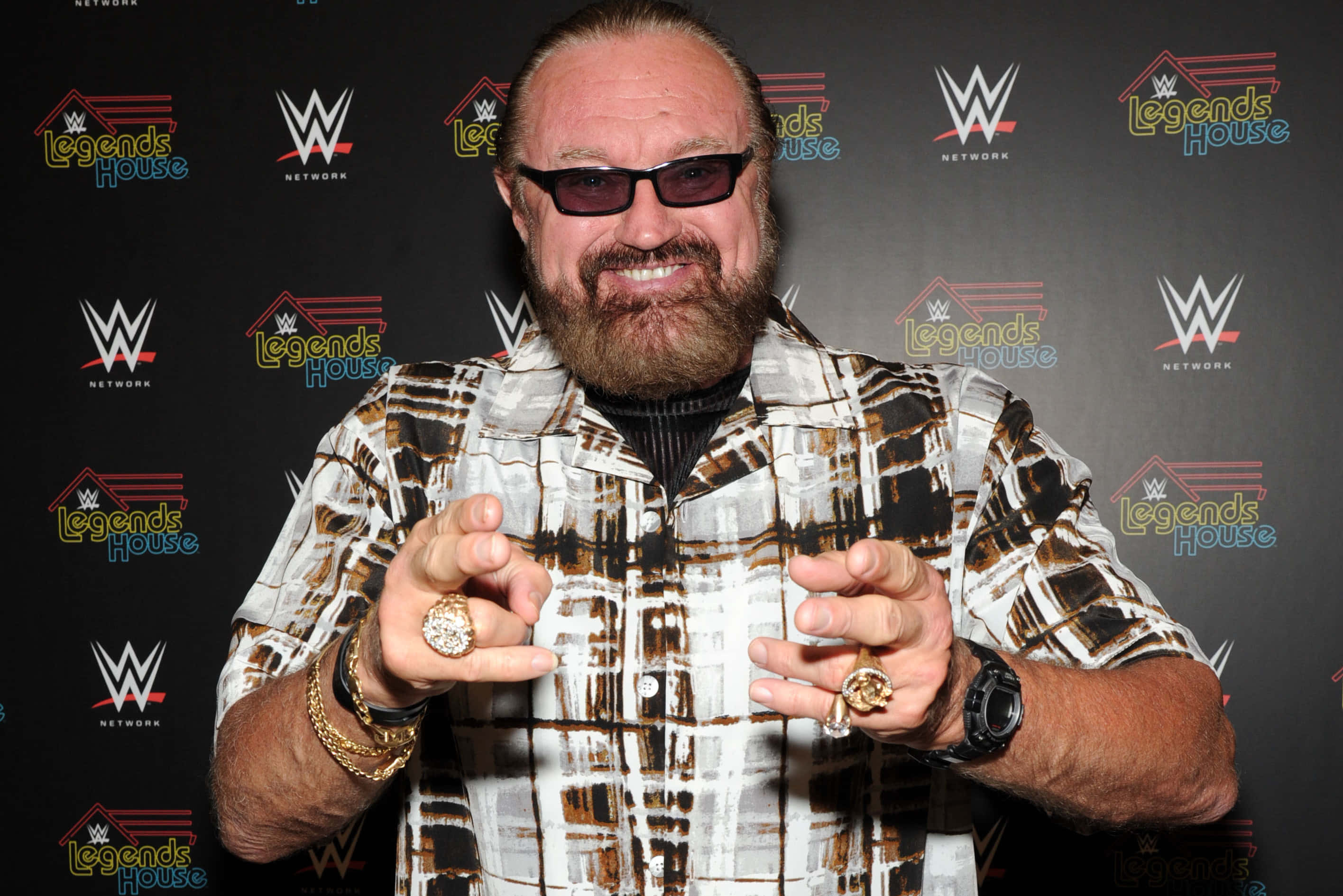 WWE 桌布 壁紙 — 一位留鬍鬚、戴太陽眼鏡、身穿圖案襯衫的男子在WWE Legends House活動中展示多枚戒指。