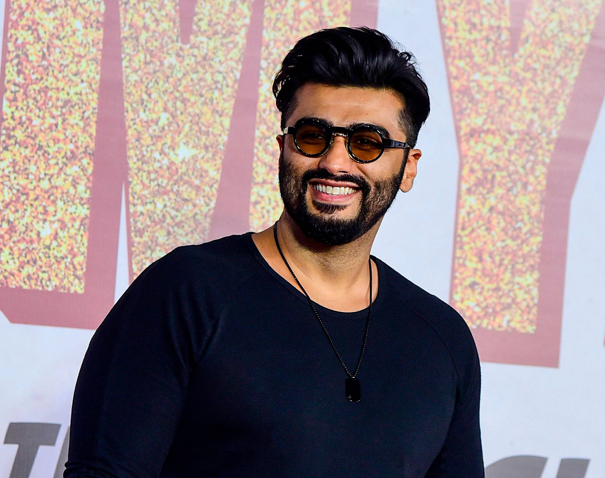Arjun Kapoor 桌布 壁紙 — 一位留鬍鬚、戴太陽眼鏡的微笑男士，在閃爍的背景前自信地擺姿勢。