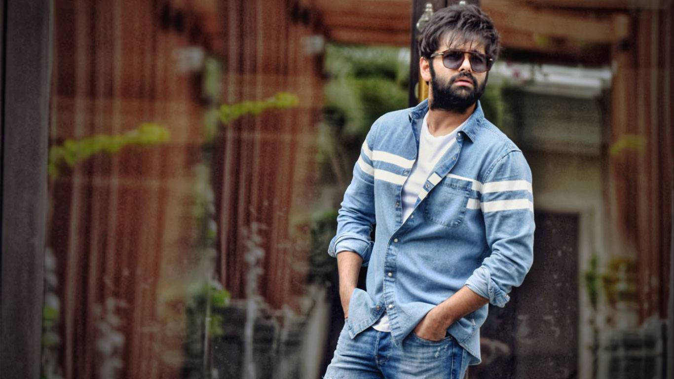 Ram Pothineni 桌布 壁紙 — 一位留鬍鬚、戴太陽眼鏡的時尚男士，穿著牛仔襯衫和牛仔褲，自信地站在模糊的城市背景前。