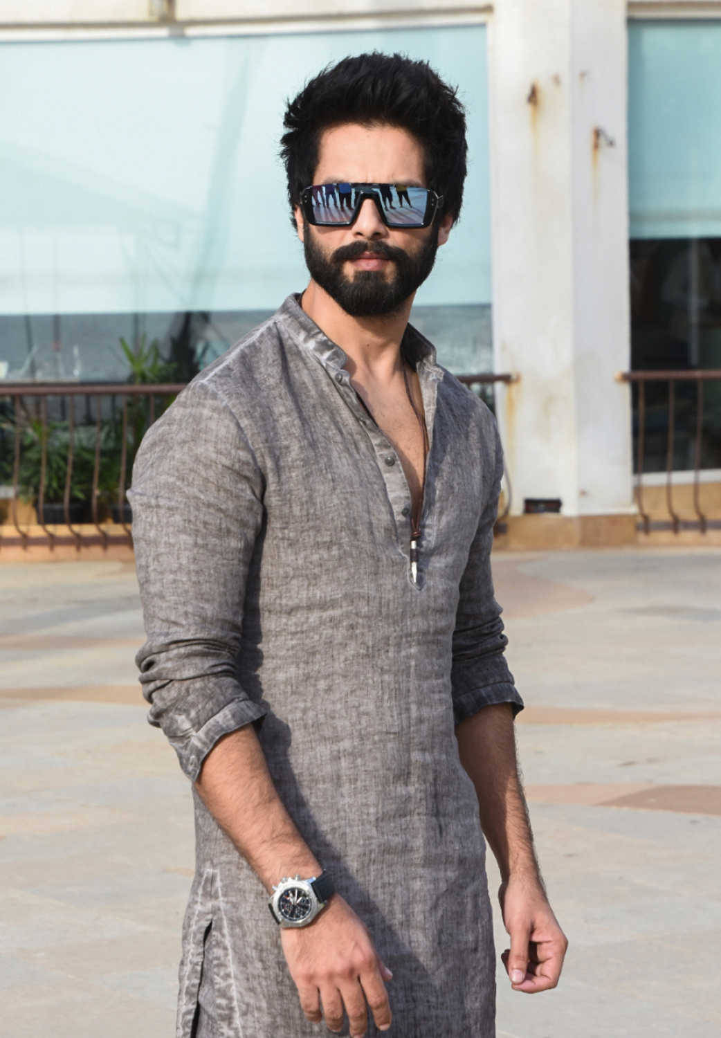 Shahid Kapoor 桌布 壁紙 — 一位留鬍鬚、戴太陽眼鏡的時尚男士，自信地穿著灰色庫爾塔，以現代建築為背景。