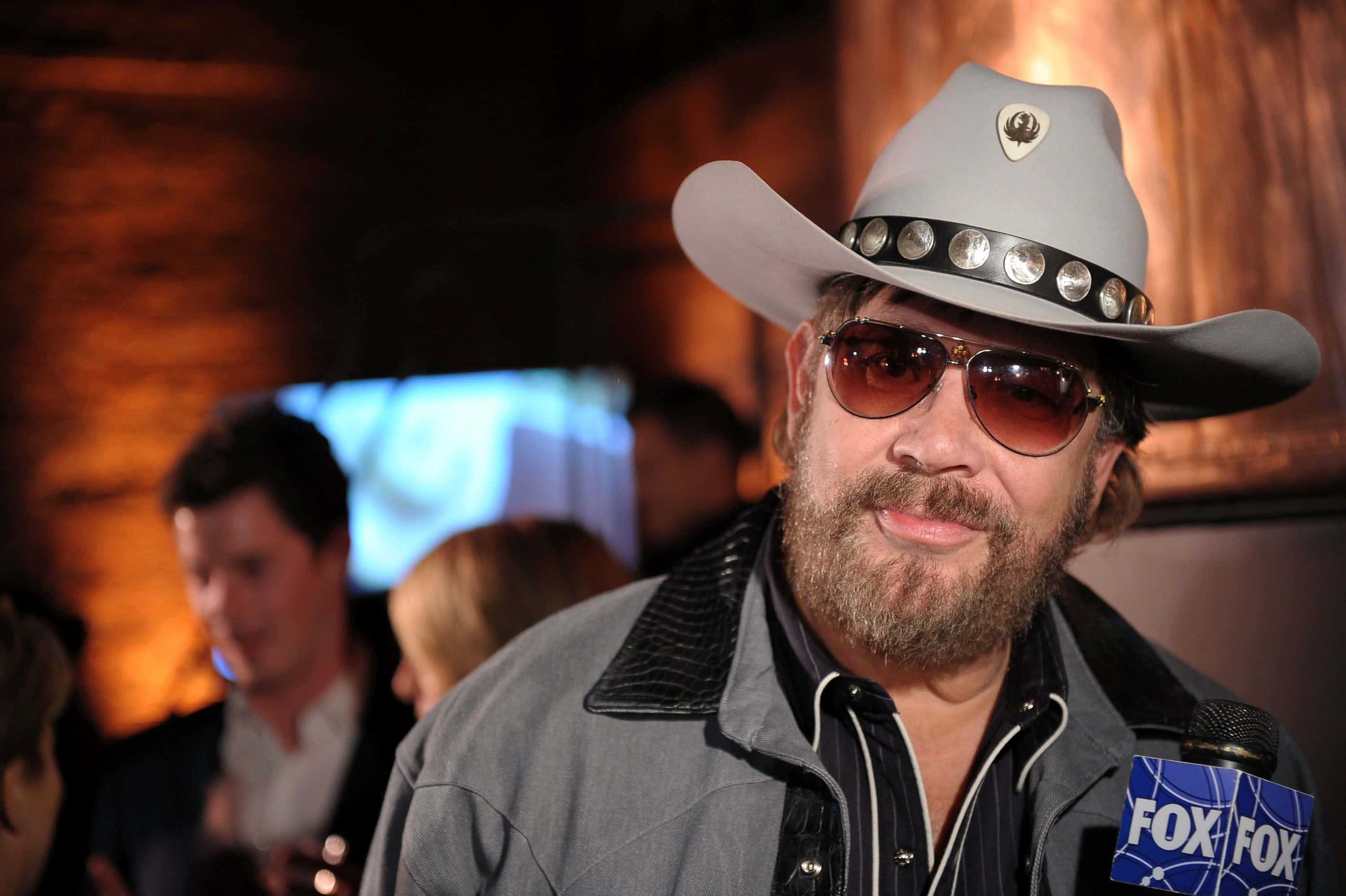 Hank Williams Jr 桌布 壁紙 — 一位留鬍鬚、戴太陽眼鏡的男子，戴著一頂帶有銀色鉚釘的灰色牛仔帽，在燈光昏暗的活動中對著FOX麥克風說話。