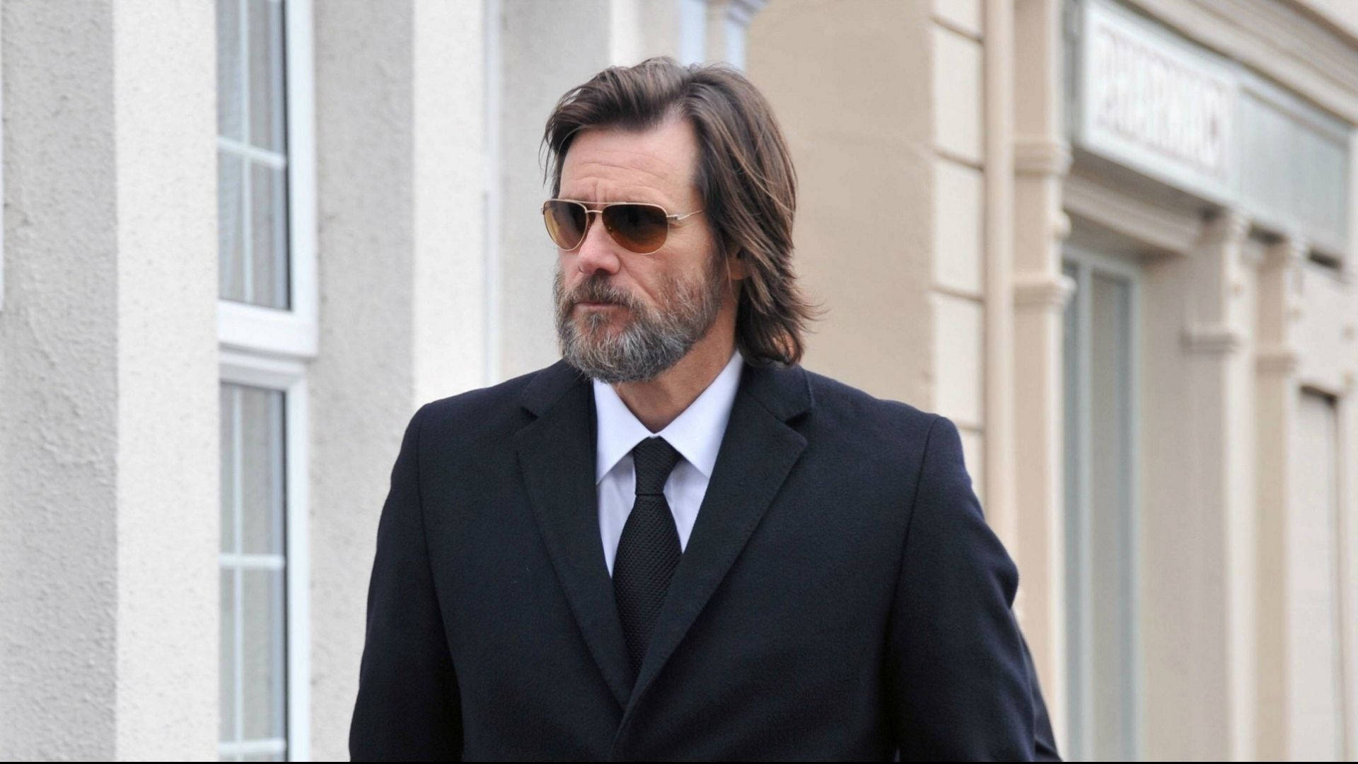 Jim Carrey 桌布 壁紙 — 一位留鬍鬚、戴太陽眼鏡的男子自信地走在陽光普照的城市街道上，經過米色建築物。