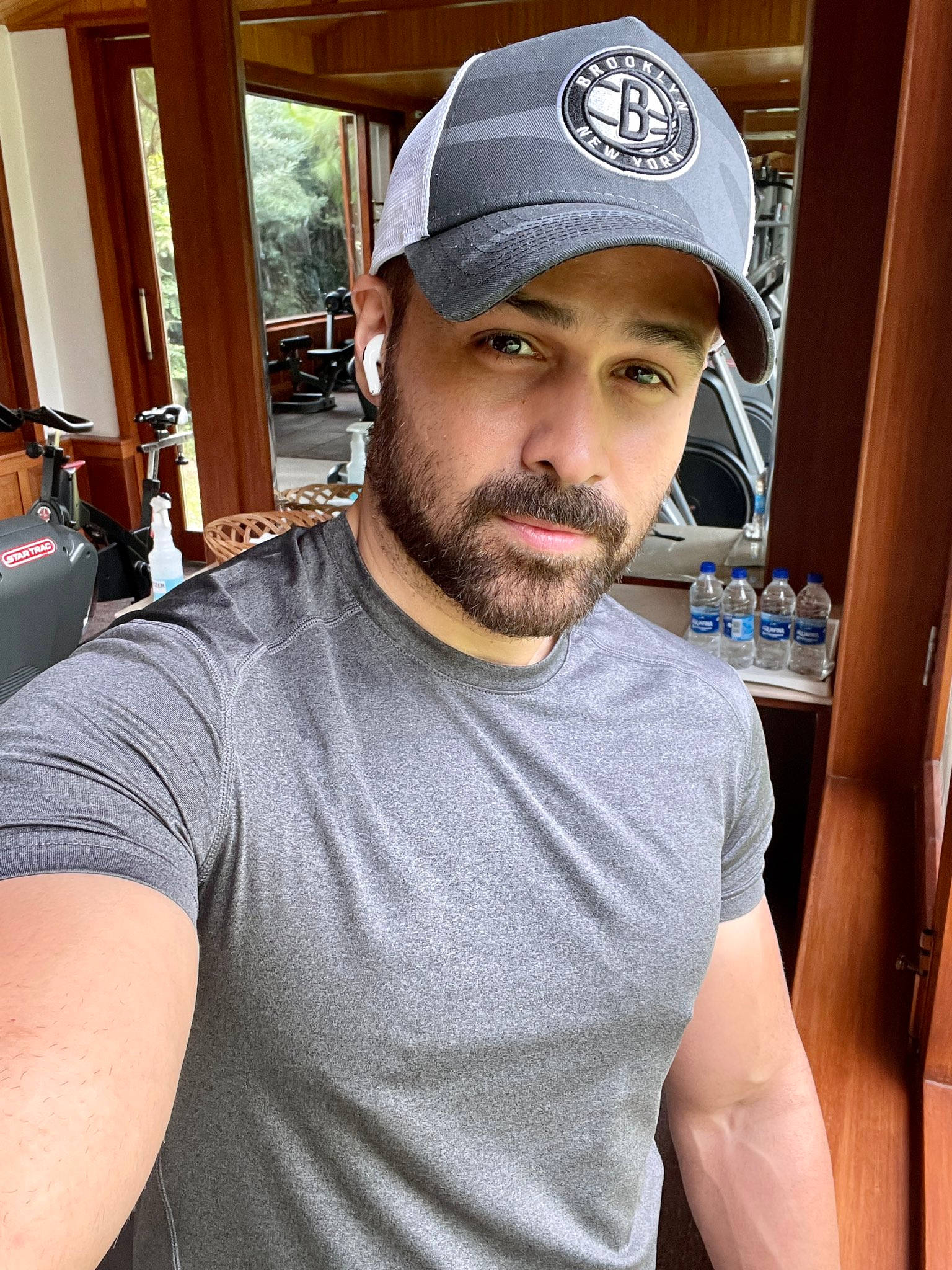 Imran 桌布 壁紙 — 一位留鬍鬚、戴灰色帽子的男人在一個有木製家具的房間裡自拍，房間內可見健身器材和水瓶。