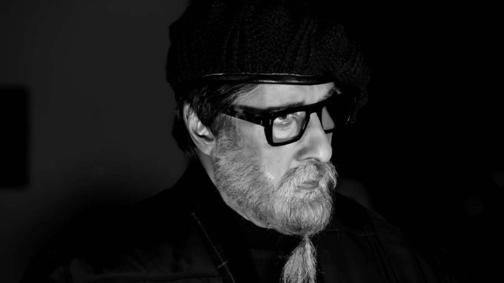 Amitabh Bachchan 桌布 壁紙 — 一位留鬍鬚、戴眼鏡和貝雷帽的男士的黑白肖像，在深色背景下若有所思地凝視。