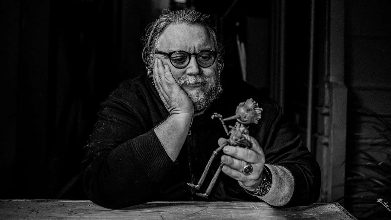 Guillermo Del Toro's Pinocchio 桌布 壁紙 — 一位留鬍鬚、戴眼鏡的男人在昏暗的房間裡，將頭靠在手上，同時拿著一個小公仔。