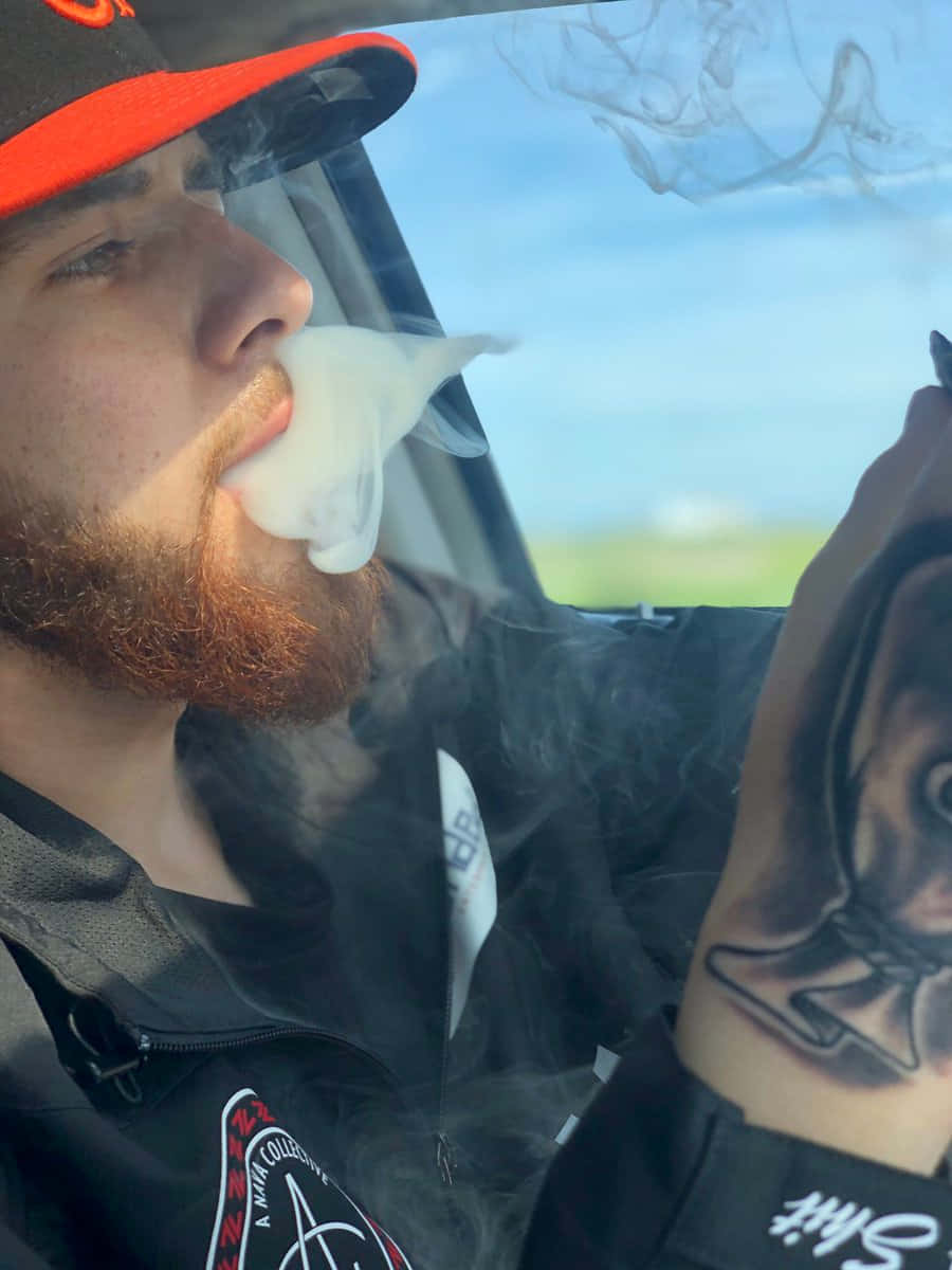 墨西哥 桌布 壁紙 — 一位留鬍鬚、戴紅帽的男人坐在車內，露出有刺青的手臂，呼出濃厚的蒸氣。