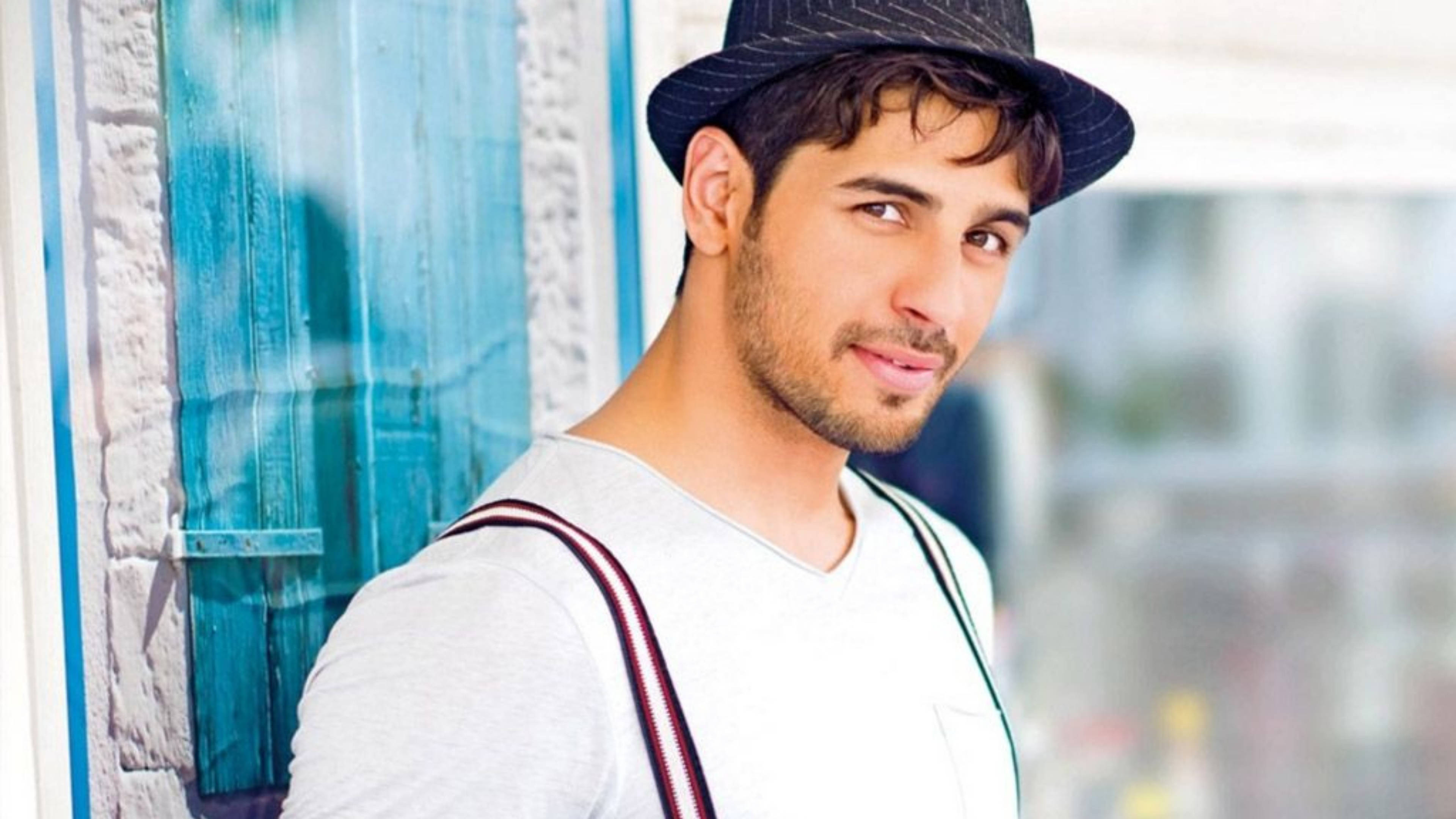 Sidharth Malhotra 桌布 壁紙 — 一位留鬍鬚、戴紳士帽的年輕男子在陽光明媚的日子裡靠著藍色木門微笑。