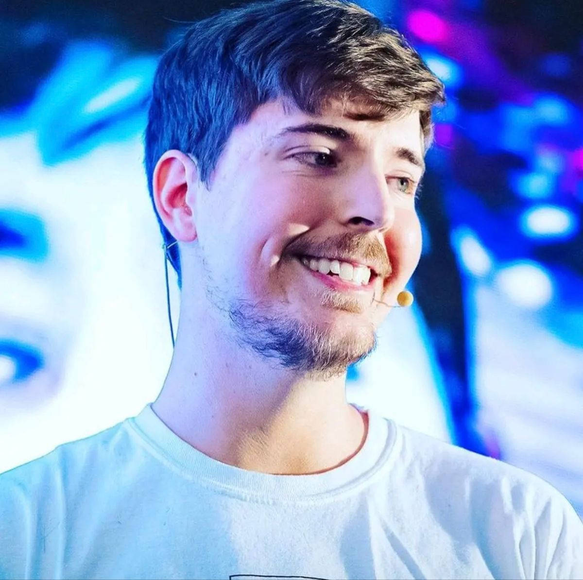 Smile 桌布 壁紙 — 一位留鬍鬚、戴耳機麥克風的微笑男士站在充滿活力的藍色和紫色燈光舞台背景前。
