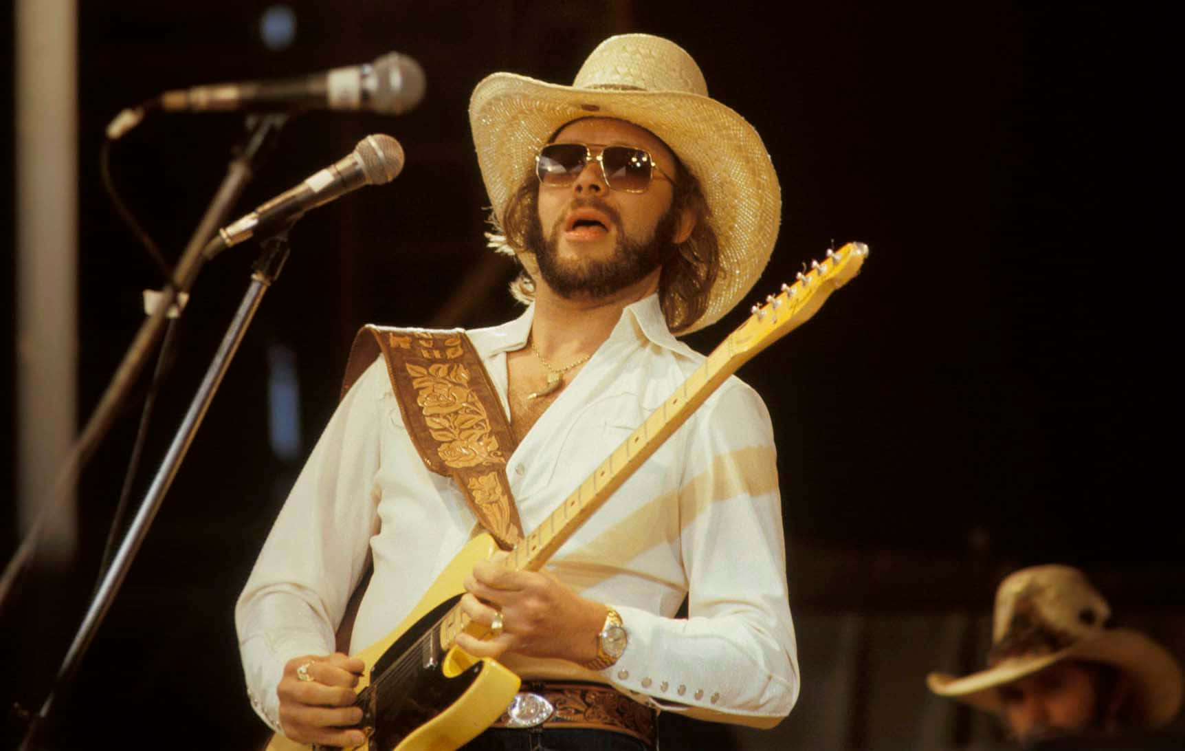 Hank Williams Jr 桌布 壁紙 — 一位留鬍鬚、戴草帽和太陽眼鏡的男子，在明亮的燈光下於舞台上熱情地彈奏一把黃色電吉他。