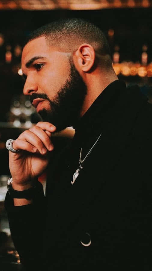 Aesthetic Drake 桌布 壁紙 — 一位留鬍鬚、戴鑽石戒指的男子，在昏暗的酒吧裡用手托著下巴，身後有溫暖的散景燈光。