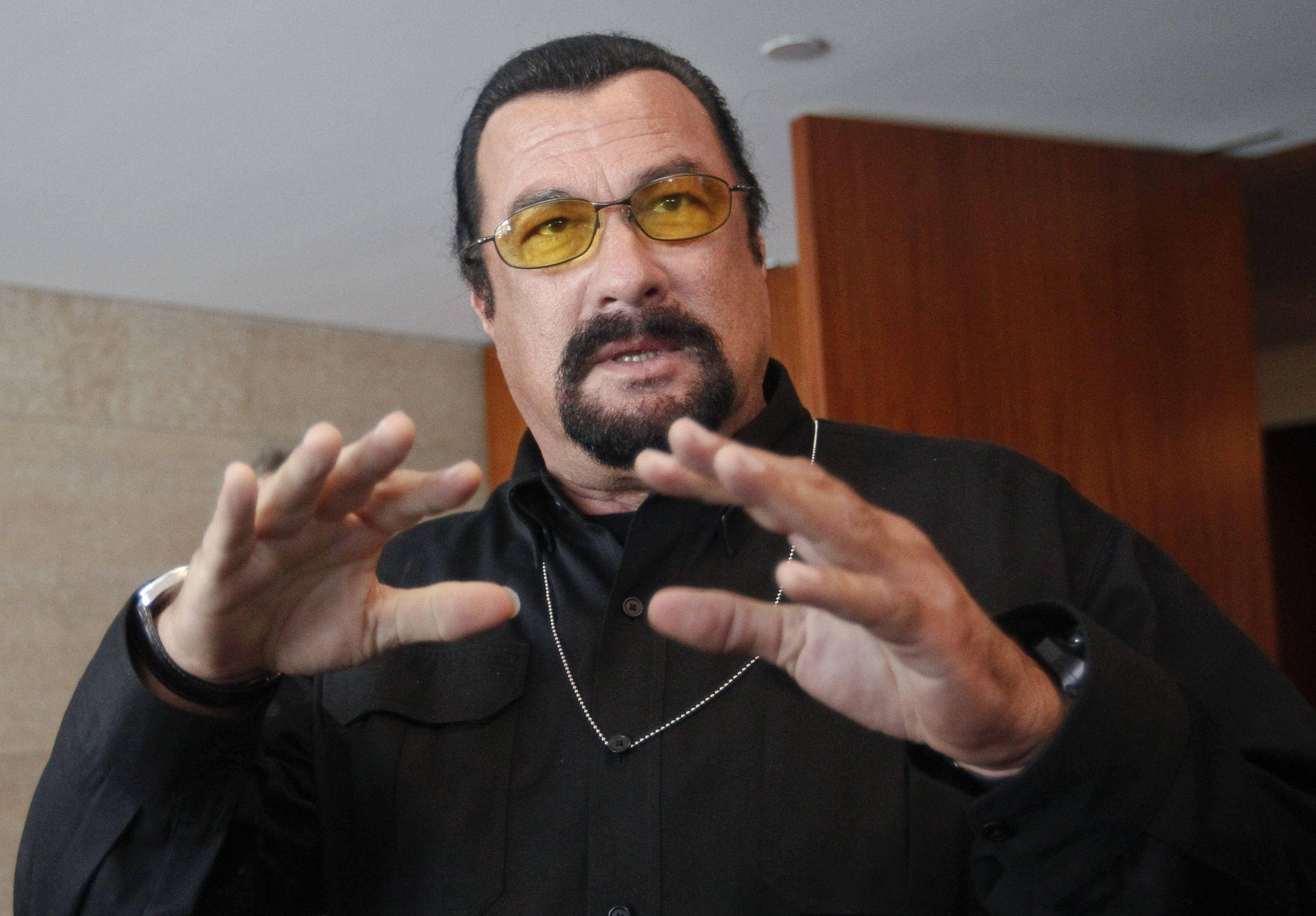 Steven Seagal 桌布 壁紙 — 一位留鬍鬚、戴黃色太陽眼鏡的男子，穿著黑色襯衫在室內生動地做手勢。