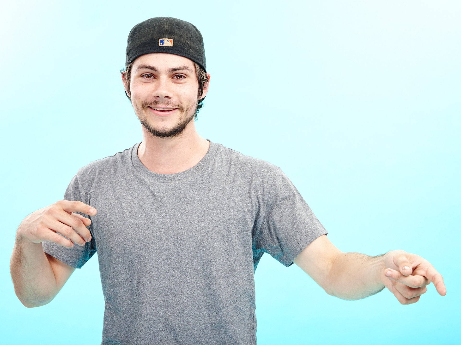 dylan 桌布 壁紙 — 一位留鬍鬚、戴黑色帽子的年輕男子，在純淺藍色背景前微笑，雙手向外指。