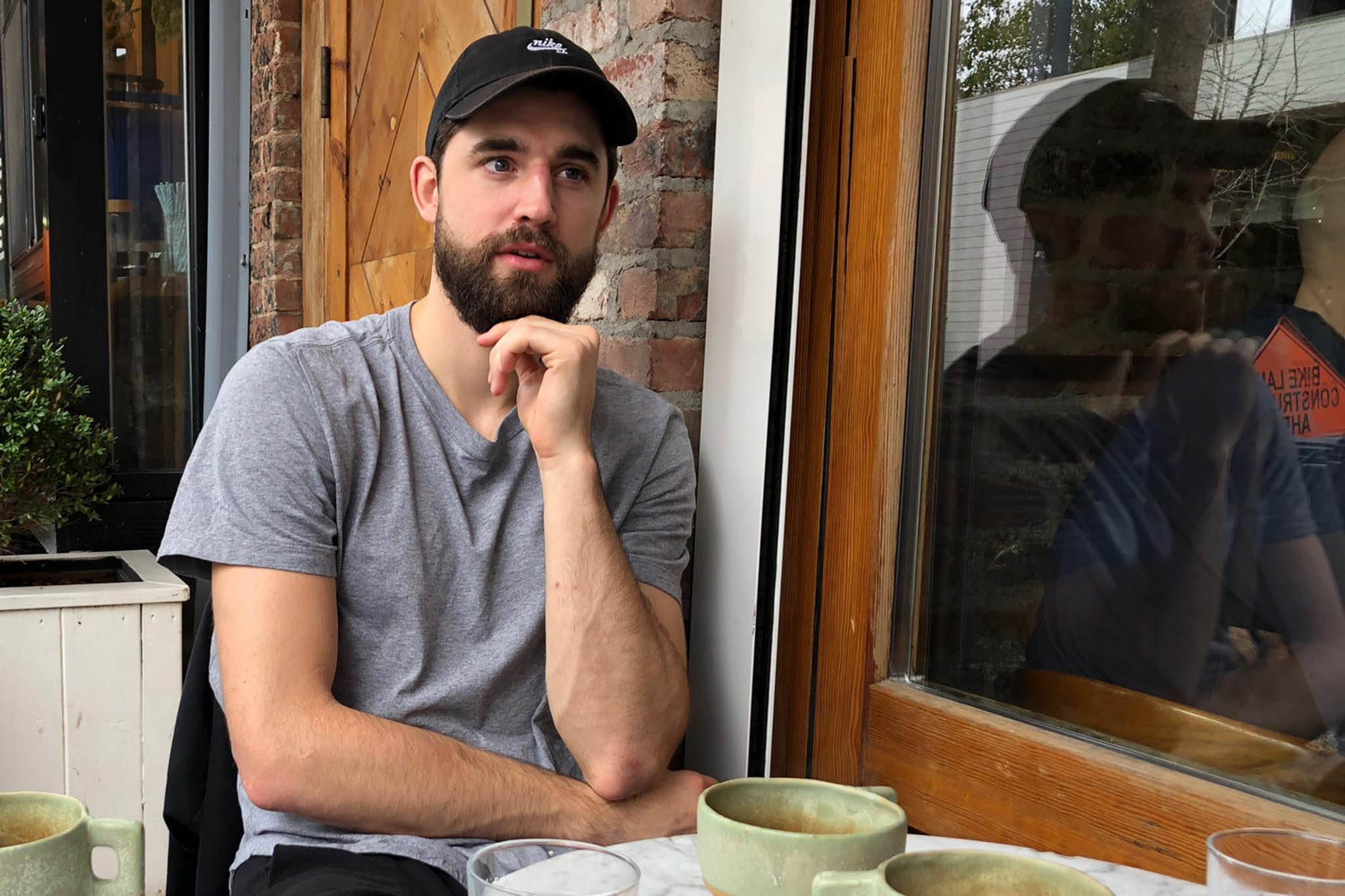 joe harris 桌布 壁紙 — 一位留鬍鬚、戴黑色帽子的男士沉思地坐在咖啡桌旁，桌上放著綠色杯子，背景是磚牆和木窗框。