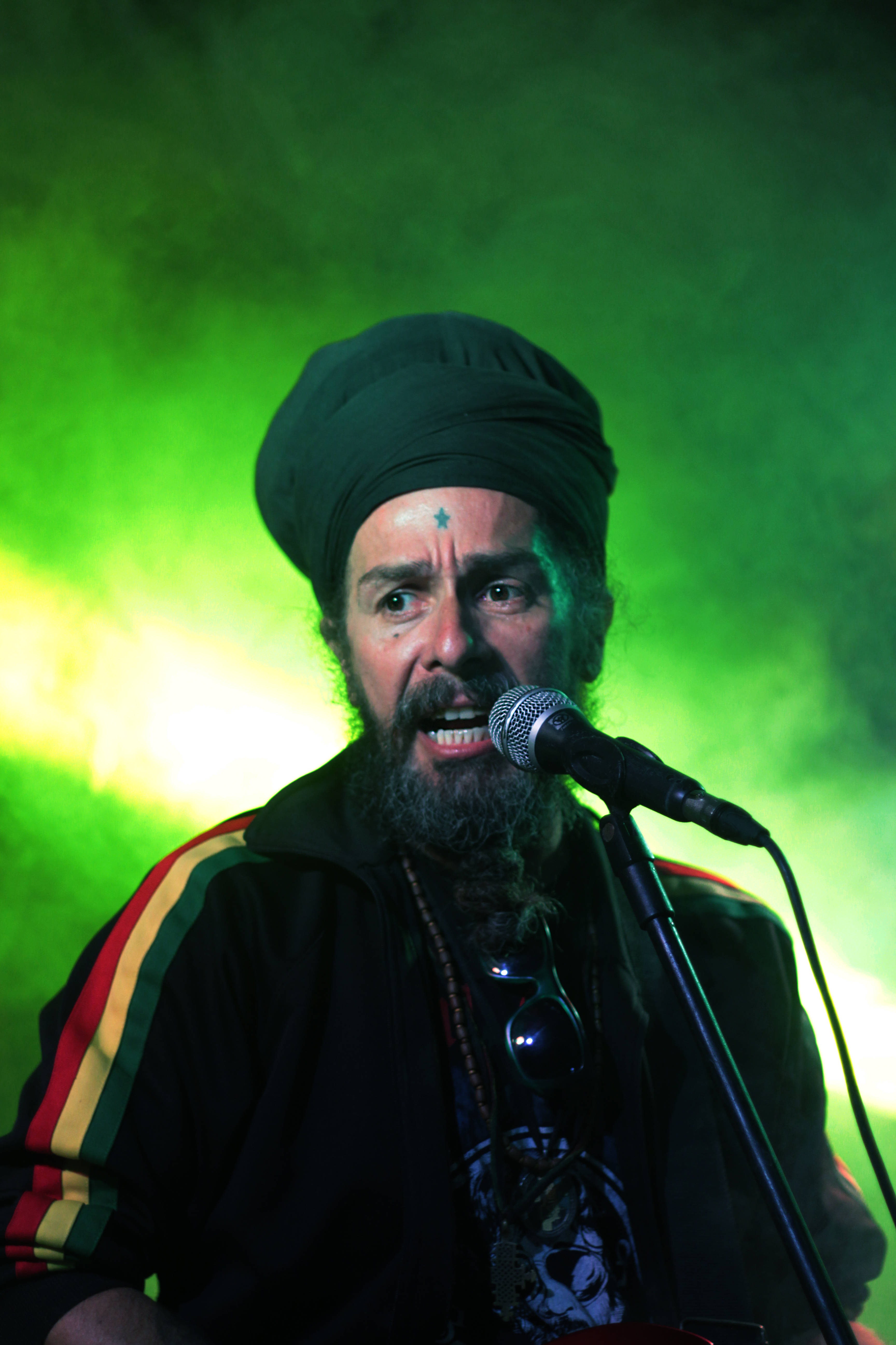 Reggae 桌布 壁紙 — 一位留鬍鬚、戴黑色頭巾、額頭有星星標記的男子，在充滿活力的綠色舞台燈光下，熱情地對著麥克風唱歌。