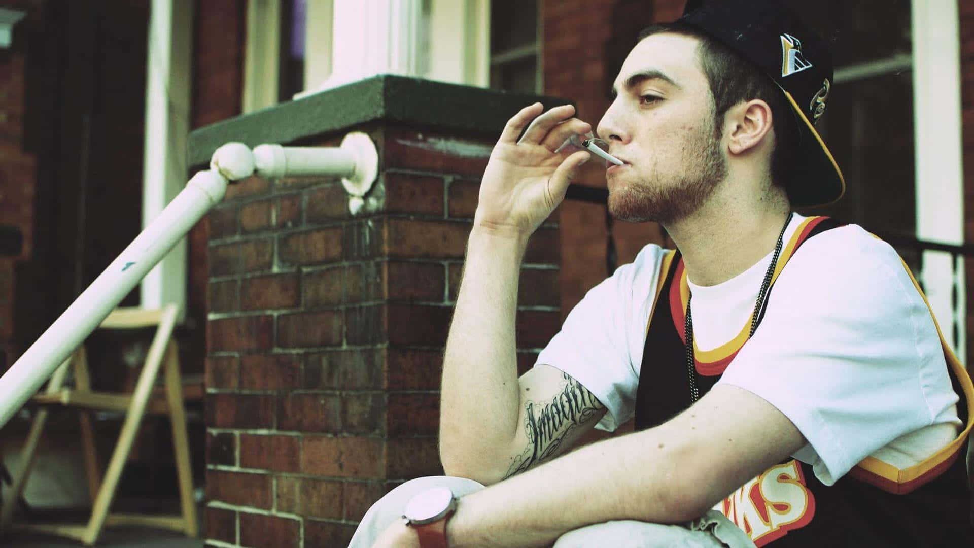 Mac Miller 桌布 壁紙 — 一位留鬍鬚、手臂有刺青的年輕男子坐在磚造建築外的台階上抽菸。