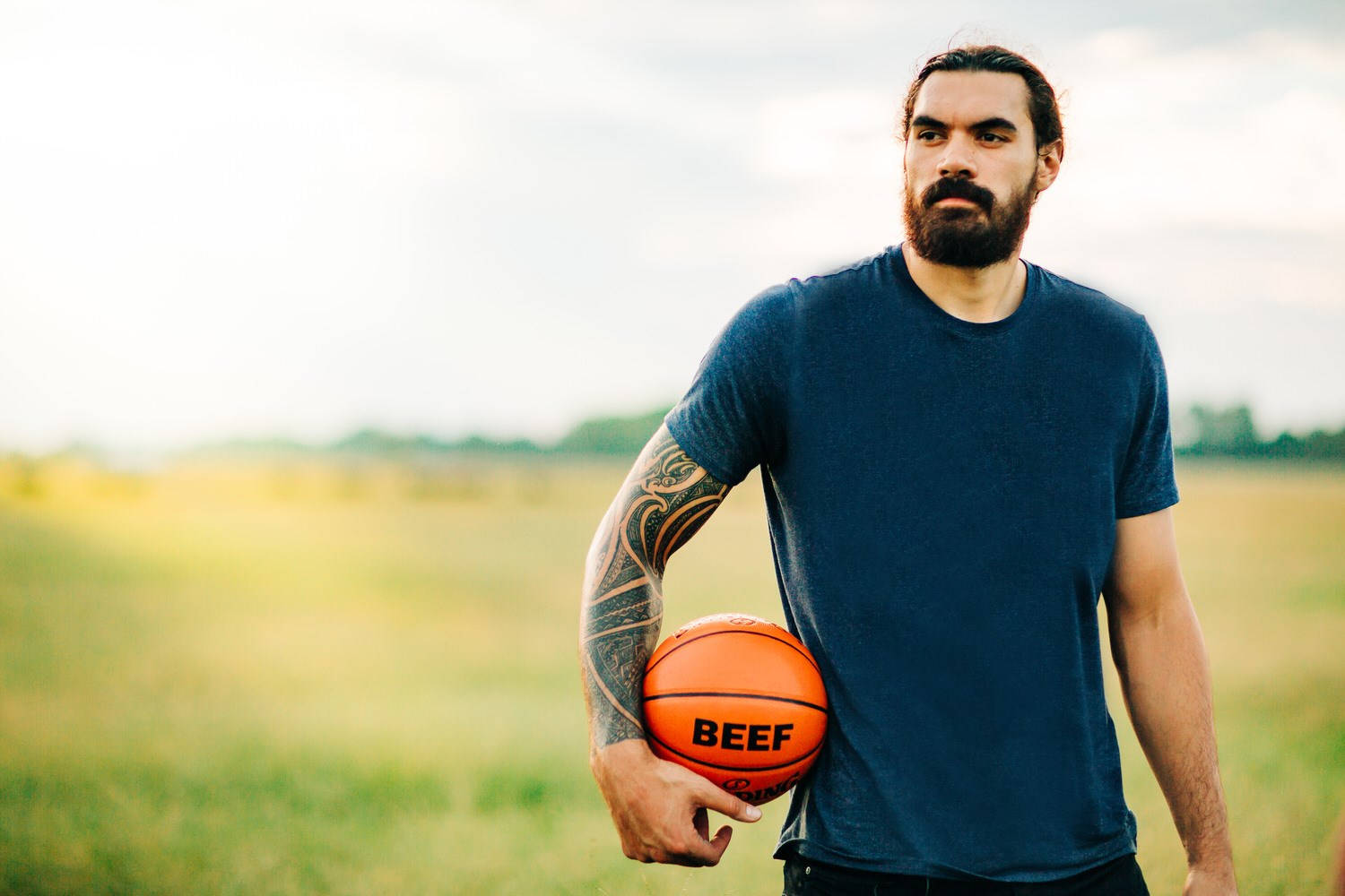 Steven Adams 桌布 壁紙 — 一位留鬍鬚、手臂有刺青的男士在陽光明媚的草地上拿著一個標有牛肉的橙色籃球。
