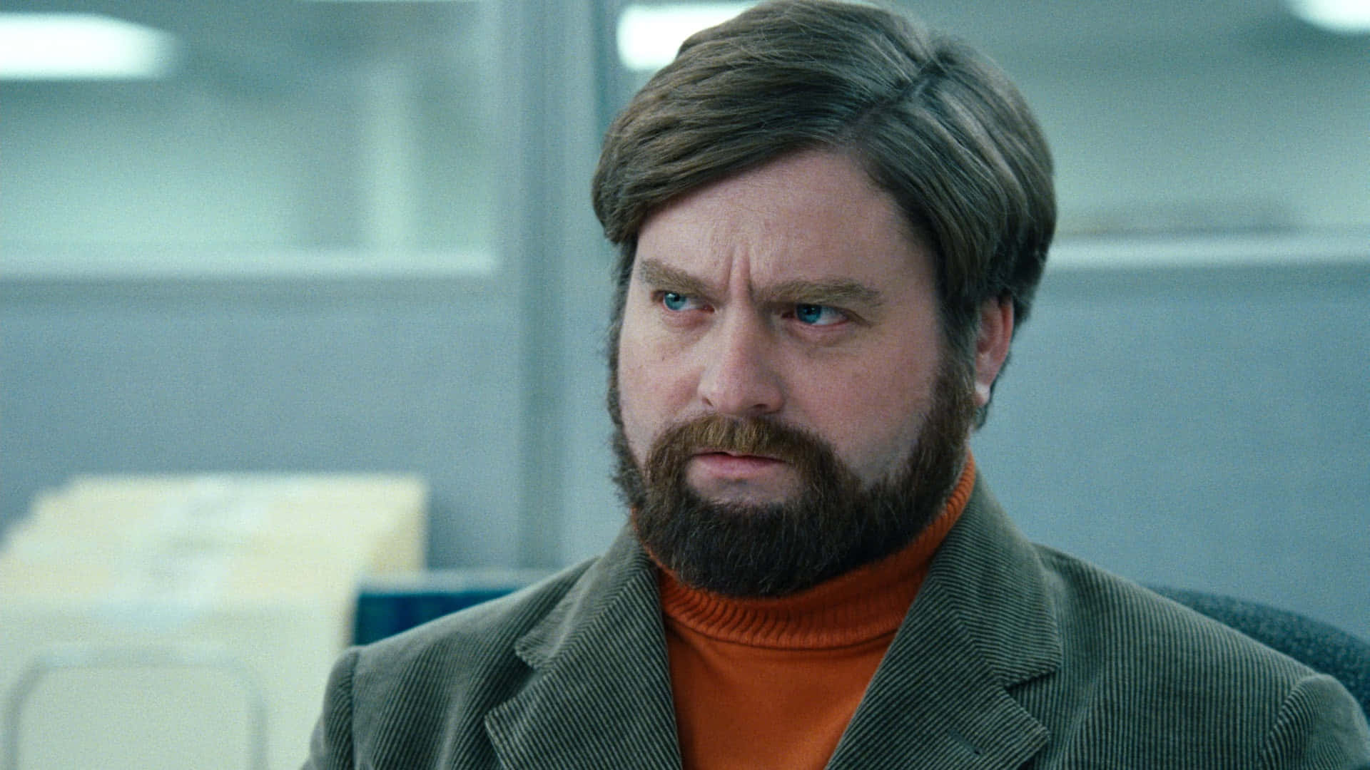 Zach Galifianakis 桌布 壁紙 — 一位留鬍鬚、擁有深邃藍眼睛的男子在辦公室環境中嚴肅地凝視，身穿橙色高領毛衣和綠色外套。