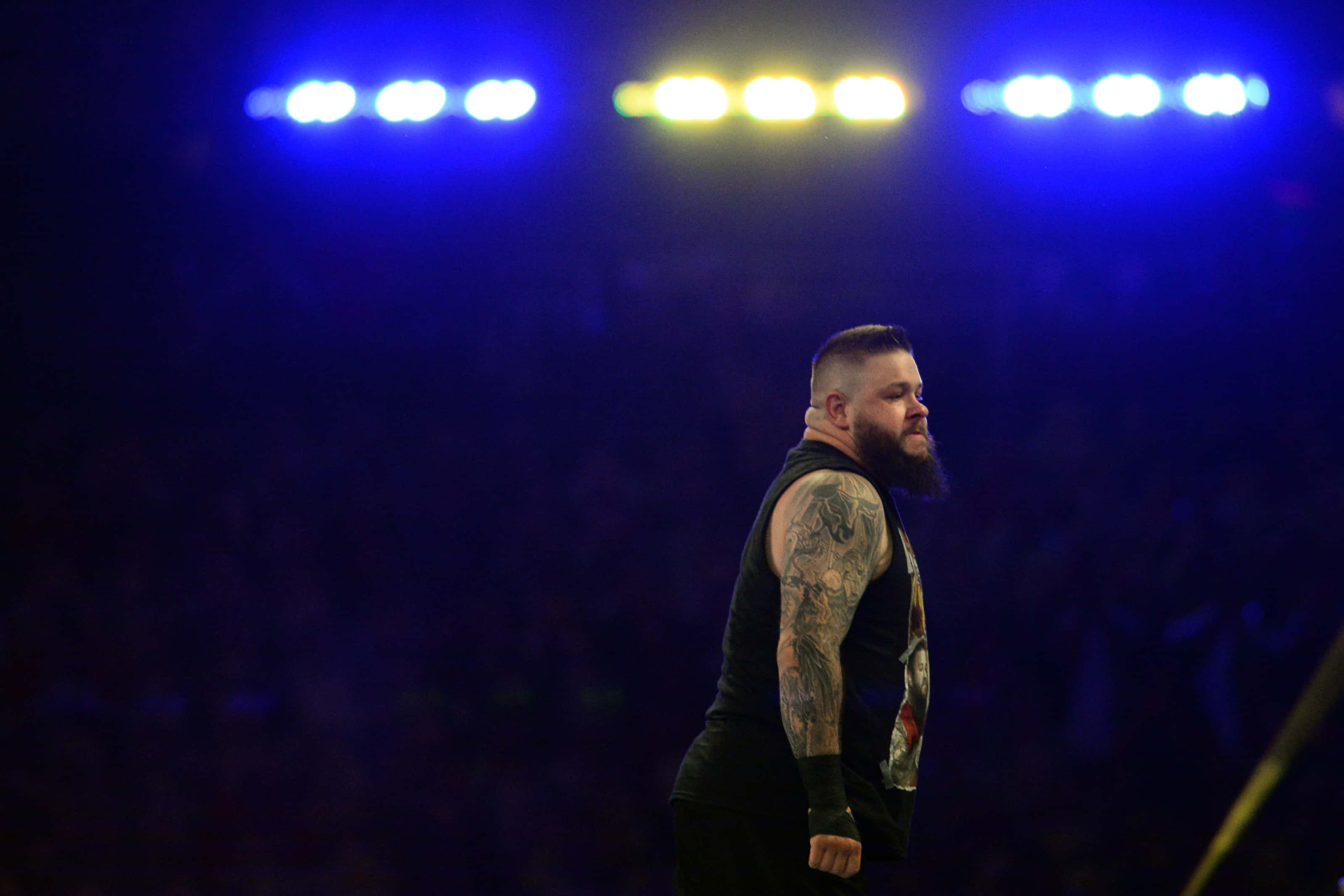 WWE 桌布 壁紙 — 一位留鬍鬚、有刺青的摔角手在明亮的藍色和黃色場館燈光下以側面站立。