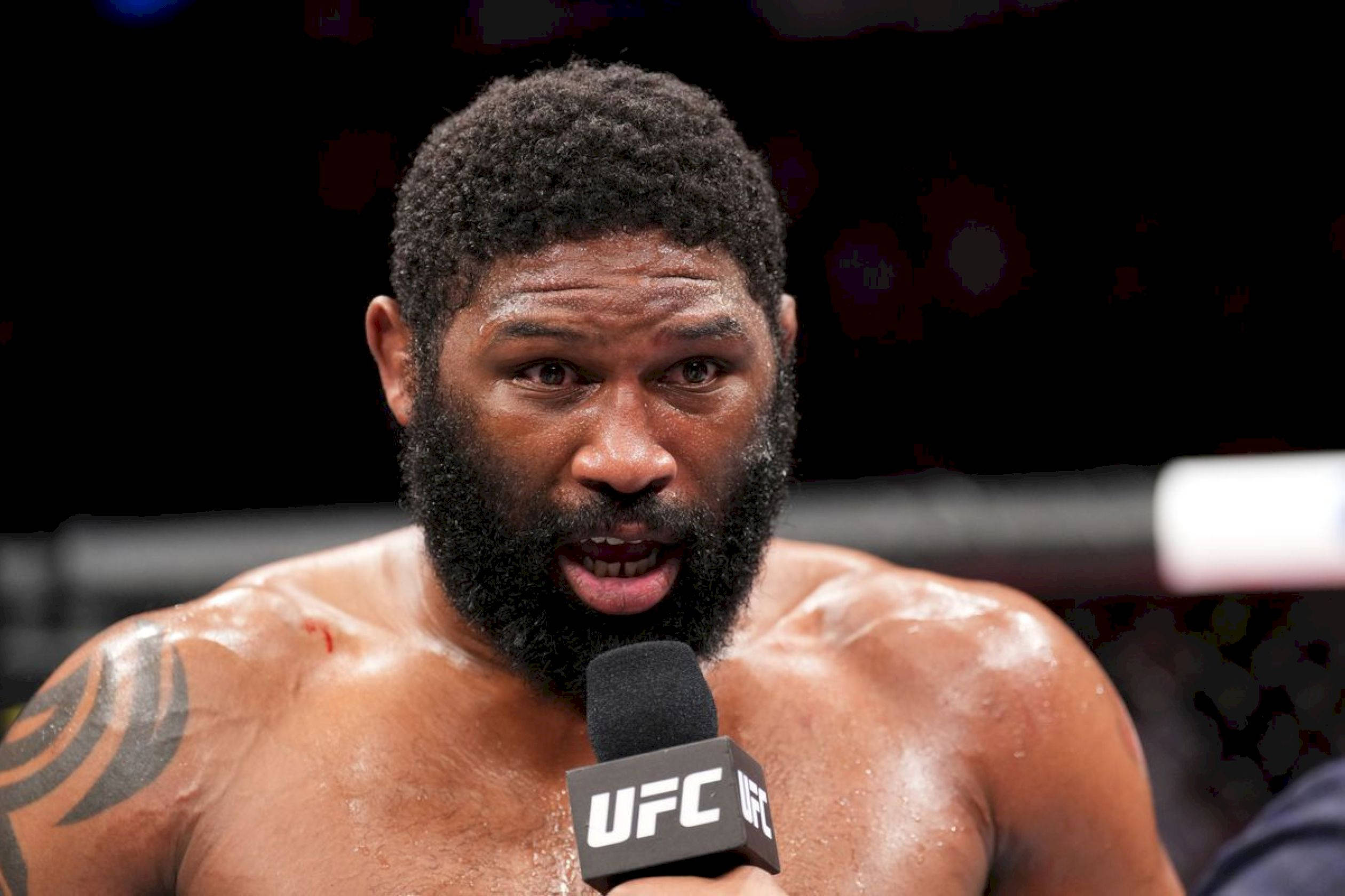 UFC 桌布 壁紙 — 一位留鬍鬚、汗流浹背的UFC拳手在明亮的場館燈光下，賽後對著麥克風說話。
