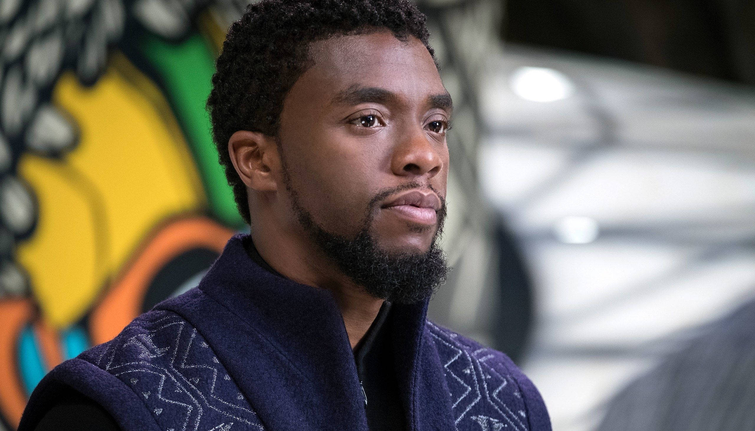 Chadwick 桌布 壁紙 — 一位留鬍鬚、穿著圖案背心的男士，在色彩繽紛的塗鴉背景下若有所思地凝視。