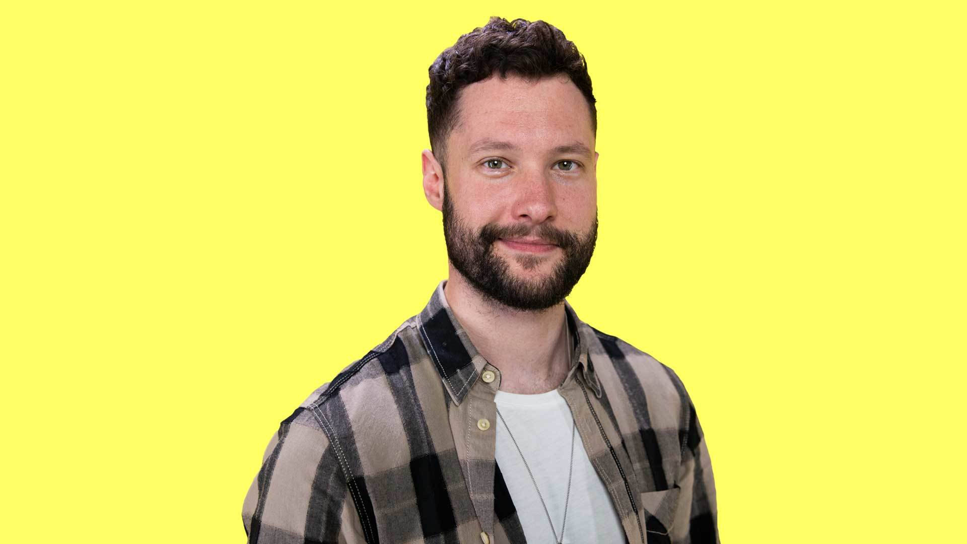 Calum Scott 桌布 壁紙 — 一位留鬍鬚、穿著格紋襯衫的男人在明亮的黃色背景前微笑。