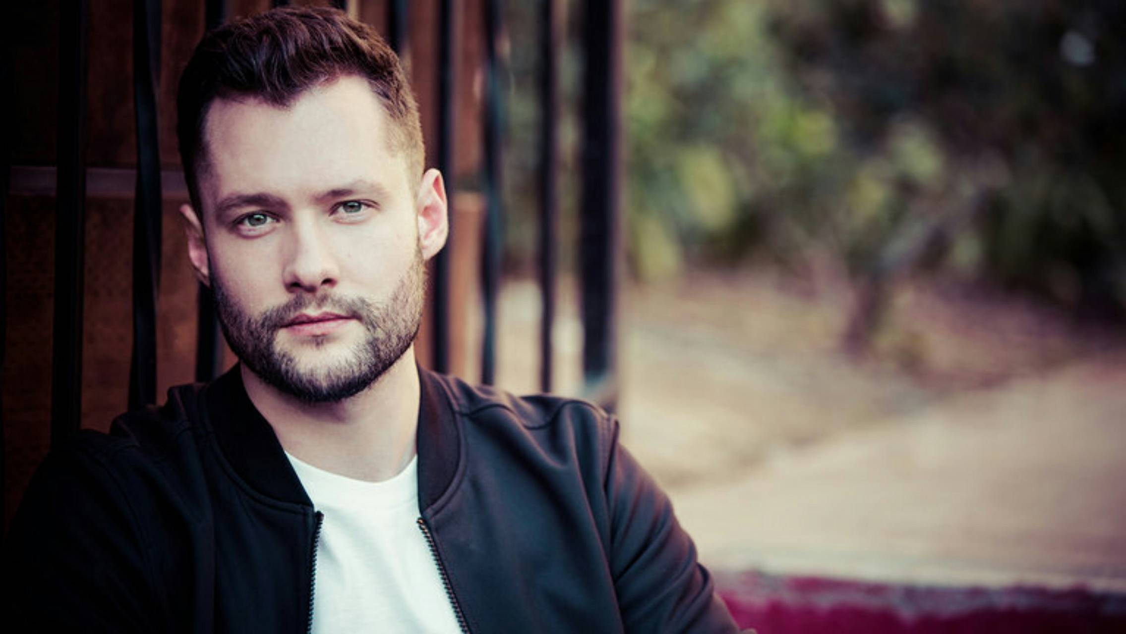 Calum Scott 桌布 壁紙 — 一位留鬍鬚、綠色眼睛的男人，身穿黑色外套內搭白色T恤，在模糊的戶外背景下直視鏡頭。