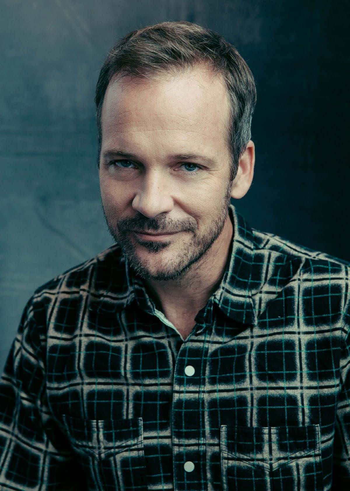 Peter Sarsgaard 桌布 壁紙 — 一位留鬍鬚、藍眼睛的男人，穿著格紋襯衫，在深色紋理背景前溫柔微笑。