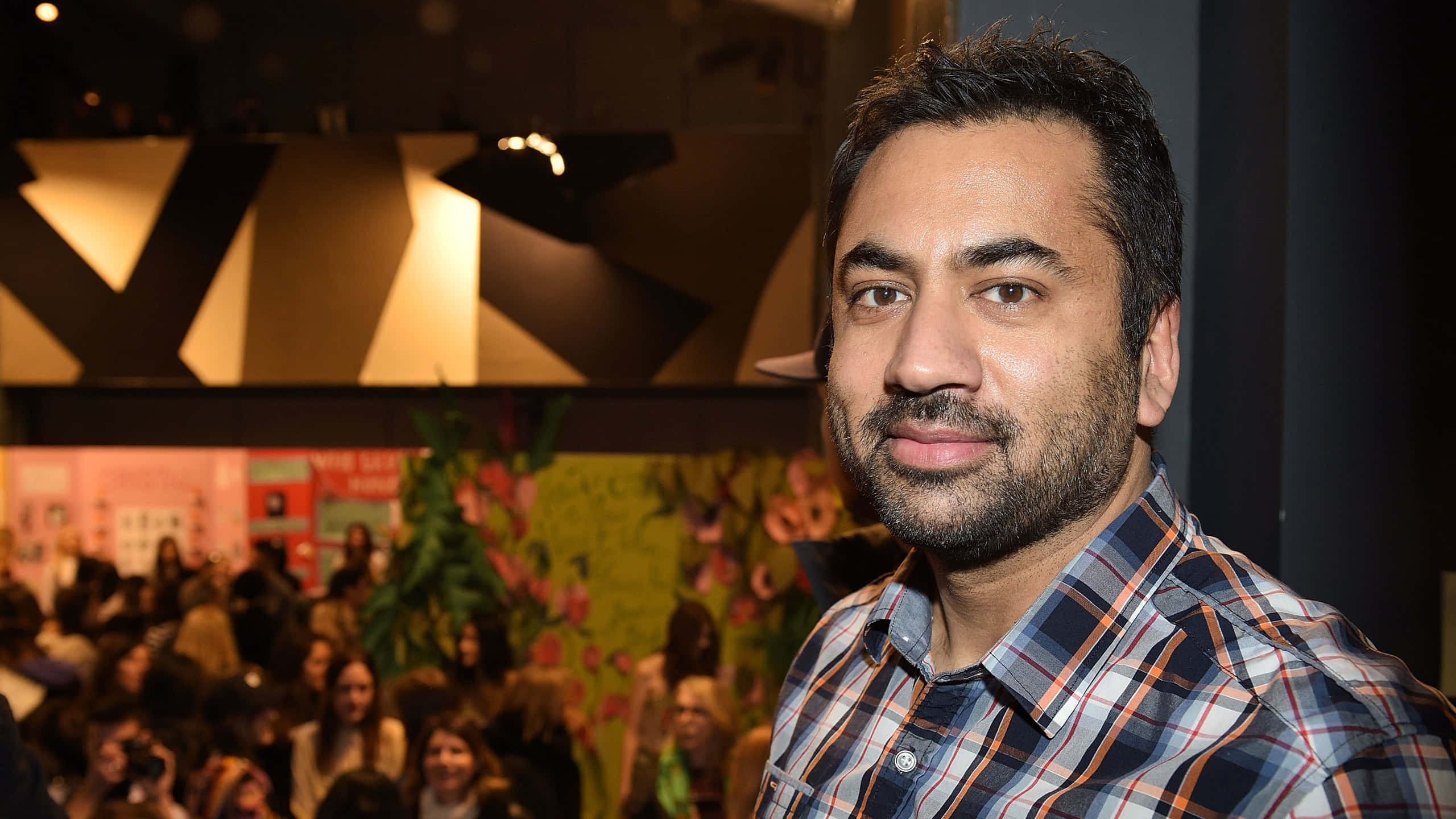 Kal Penn 桌布 壁紙 — 一位留鬍鬚、身穿格紋襯衫的男士在燈光溫暖的室內活動中看向鏡頭，背後有模糊的人群和花卉裝飾。