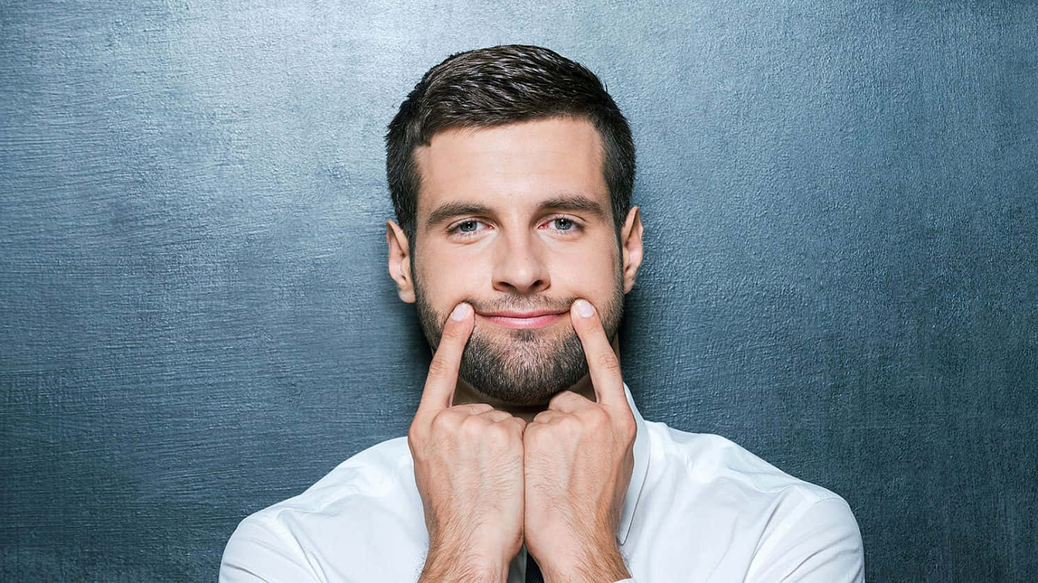 Fake Smile 桌布 壁紙 — 一位留鬍鬚、身穿白襯衫的男子微笑著，將臉頰貼在帶有紋理的藍灰色牆上。