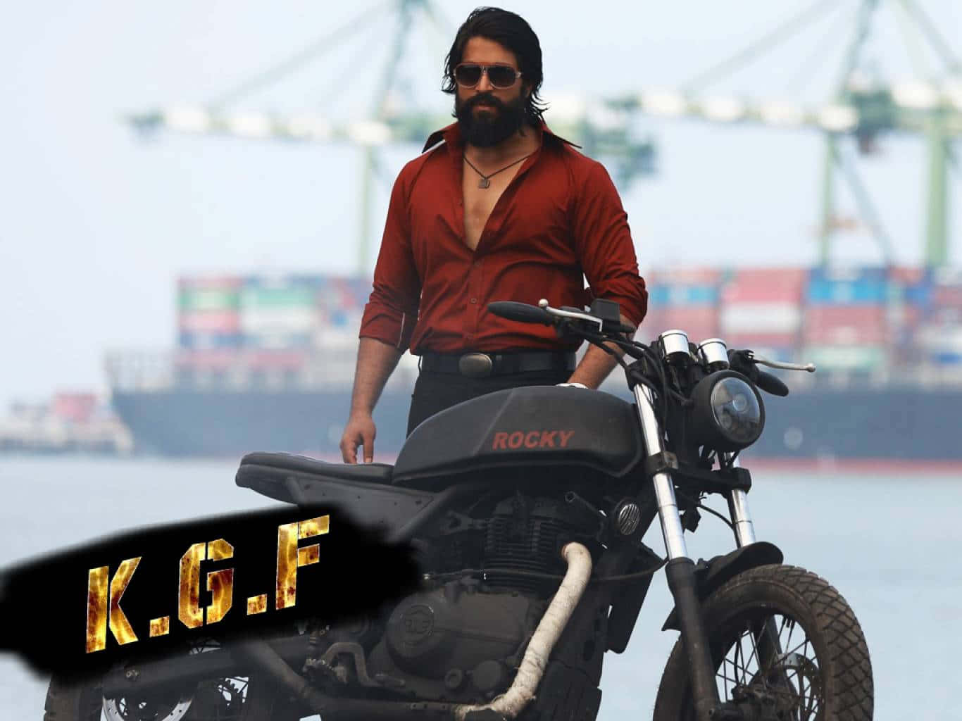 Kgf 桌布 壁紙 — 一位留鬍鬚、身穿紅衫的男子自信地站在一輛黑色機車旁，機車油箱上寫有「ROCKY」，附近是貨船和起重機的港口。