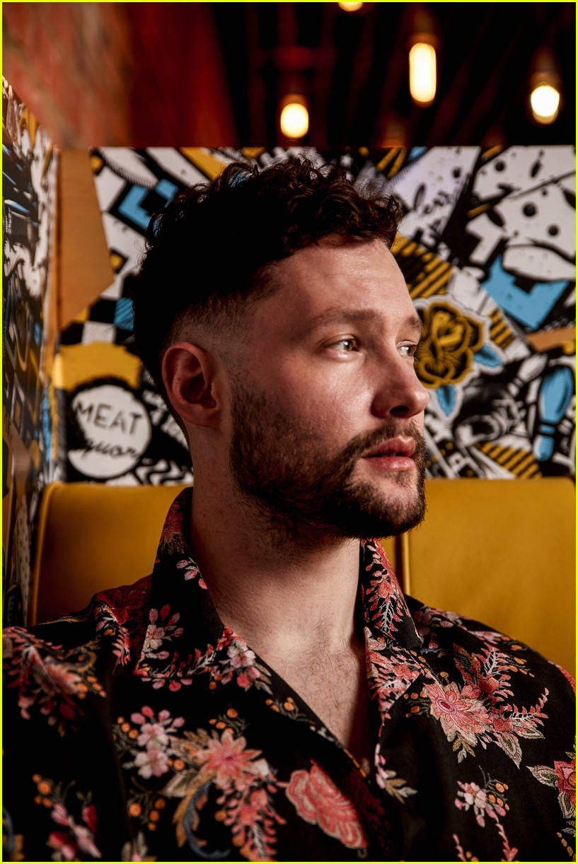Calum Scott 桌布 壁紙 — 一位留鬍鬚、身穿花卉圖案襯衫的男子，在一個光線溫暖、帶有漫畫風格牆藝的房間裡若有所思地凝視著。