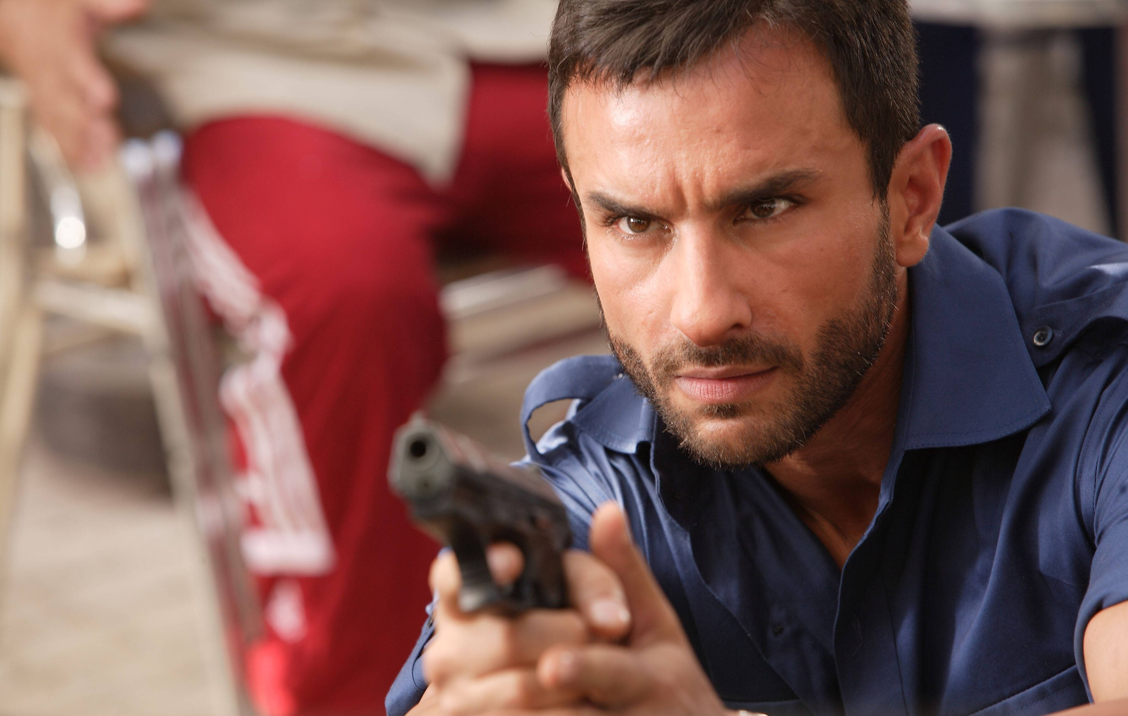 Saif Ali Khan 桌布 壁紙 — 一位留鬍鬚、身穿藍色襯衫的男子，在模糊的室內環境中，專注地瞄準手槍。