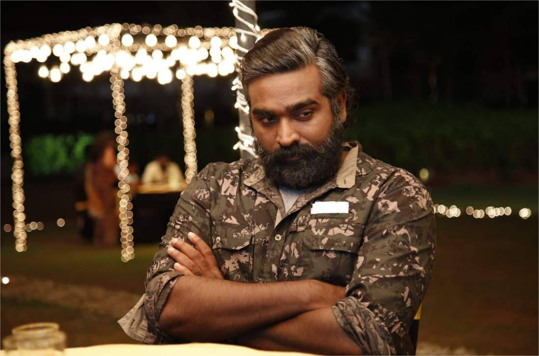 Vijay Sethupathi HD 桌布 壁紙 — 一位留鬍鬚、身穿迷彩襯衫的男子在夜晚交叉雙臂坐在發光的串燈下。