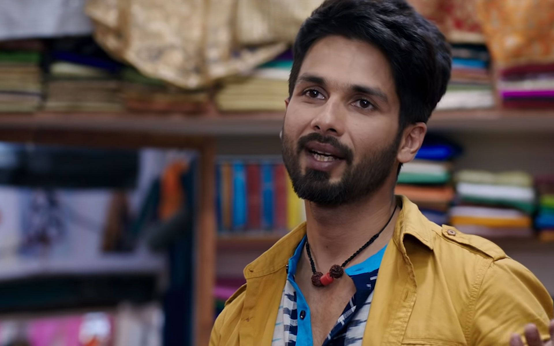 Shahid Kapoor 桌布 壁紙 — 一位留鬍鬚、身穿黃色外套的男子在色彩繽紛的布料店裡溫暖地微笑。
