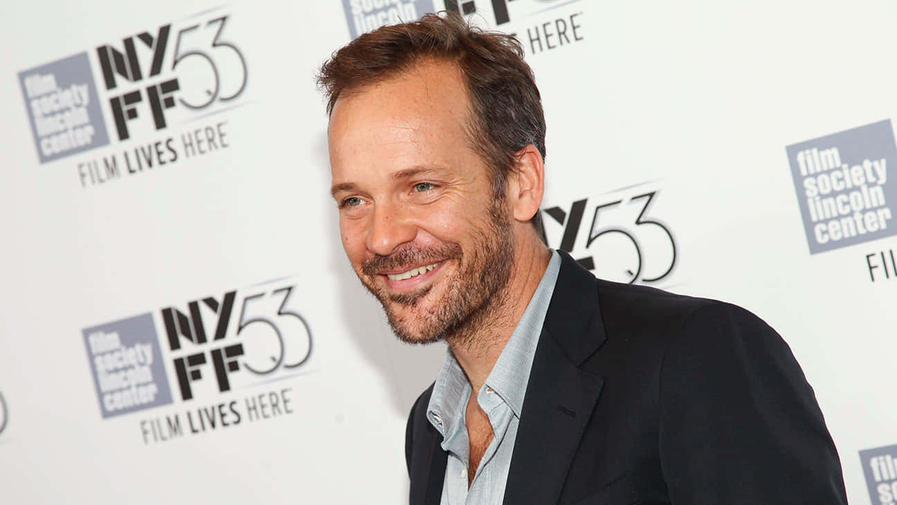 Peter Sarsgaard 桌布 壁紙 — 一位留鬍鬚、面帶微笑的男士，身穿深色西裝和淺色襯衫，在有品牌背景的電影節活動中擺姿勢。